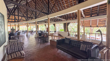 Long open lodge gallery with leather and patterned chairs, red tile floor, timber posts, and exposed thatched roof.