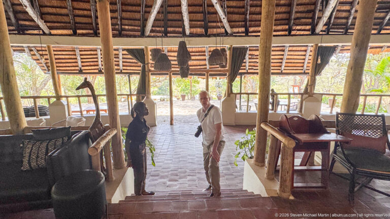 Two people talking on steps inside an open-sided lodge with timber posts, woven furniture, and hanging shades.