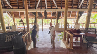 Two people talking on steps inside an open-sided lodge with timber posts, woven furniture, and hanging shades.