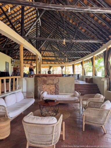 Pale wicker sofas and chairs grouped around low tables beneath exposed lodge roof trusses, with a bar behind.