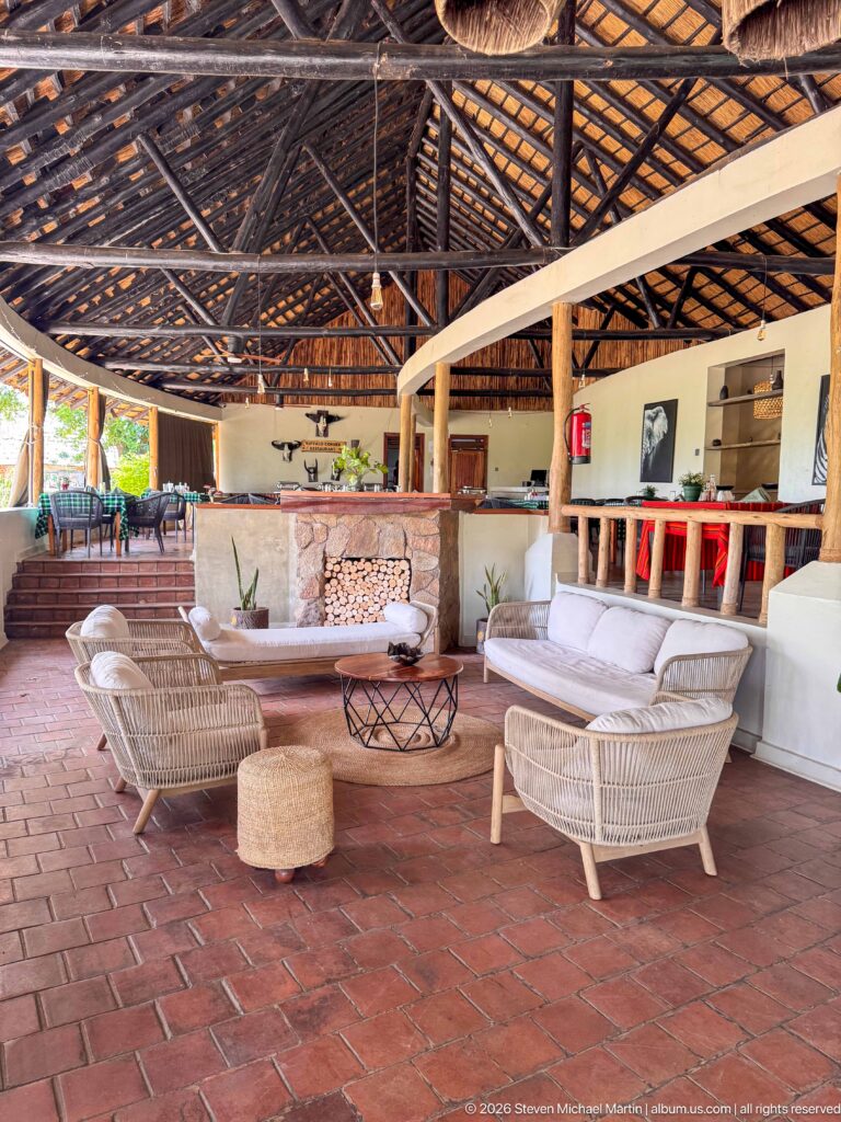 Open lodge lounge with pale wicker seating, red tile floor, raised bar, stacked firewood, and a high thatched roof.