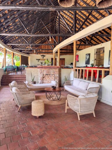 Open lodge lounge with pale wicker seating, red tile floor, raised bar, stacked firewood, and a high thatched roof.