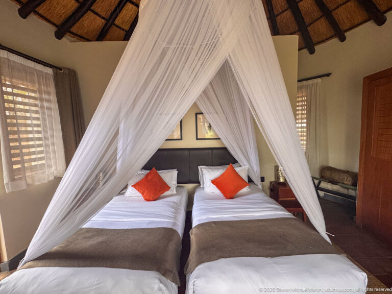 Front view of two single beds under a peaked white mosquito net, with orange cushions and louvered windows.