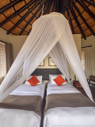 Two single beds with gray throws and orange cushions enclosed by a large white mosquito net beneath a thatched roof.