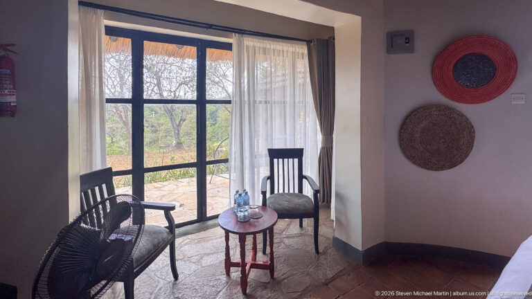 Two dark woven chairs and a small round table beside glazed doors overlooking dry woodland in a lodge room.