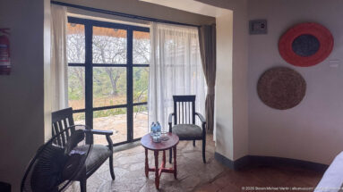 Two dark woven chairs and a small round table beside glazed doors overlooking dry woodland in a lodge room.