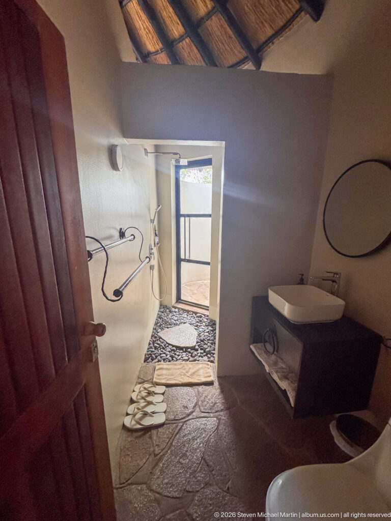 Rustic bathroom with stone floor, walk-in shower, white basin and toilet, towels, and daylight through an open doorway.