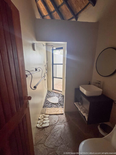 Rustic bathroom with stone floor, walk-in shower, white basin and toilet, towels, and daylight through an open doorway.
