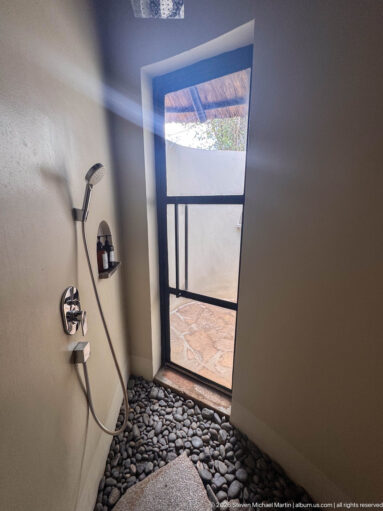 Walk-in shower with pale walls, handheld fixture, dark river-stone floor, and open doorway to a tiled exterior area.