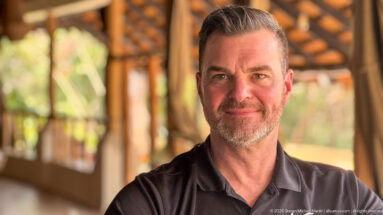Close portrait of a bearded man in a dark polo beneath a thatched lodge roof, with greenery behind.