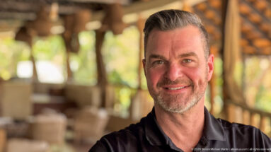 Smiling bearded man in a black polo with an open timber-and-thatch lodge softly blurred behind him.