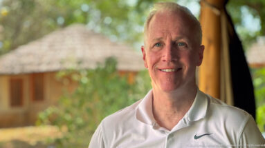 Smiling man in a white polo shirt standing in a shaded lodge with a blurred thatched building behind him.