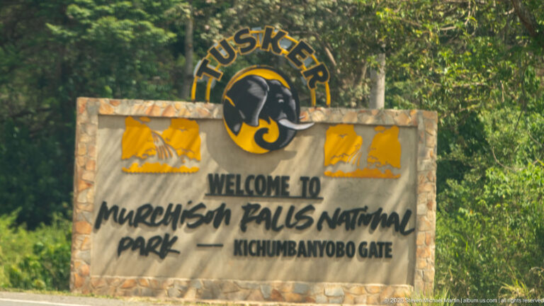 Stone sign reading “Welcome to Murchison Falls National Park — Kichumbanyobo Gate,” topped by a Tusker emblem.