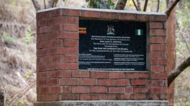 Black commemorative plaque with Ugandan and Nigerian flags mounted in a red-brick monument amid dry woodland.