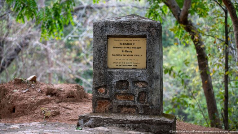 Tall stone commemorative monument with a brass plaque standing beneath trees near Murchison Falls.
