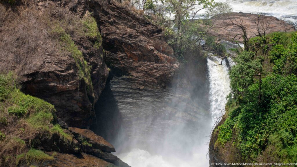 Narrow white waterfall chute descending between steep dark rock walls with green vegetation on one side.