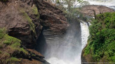 Narrow white waterfall chute descending between steep dark rock walls with green vegetation on one side.