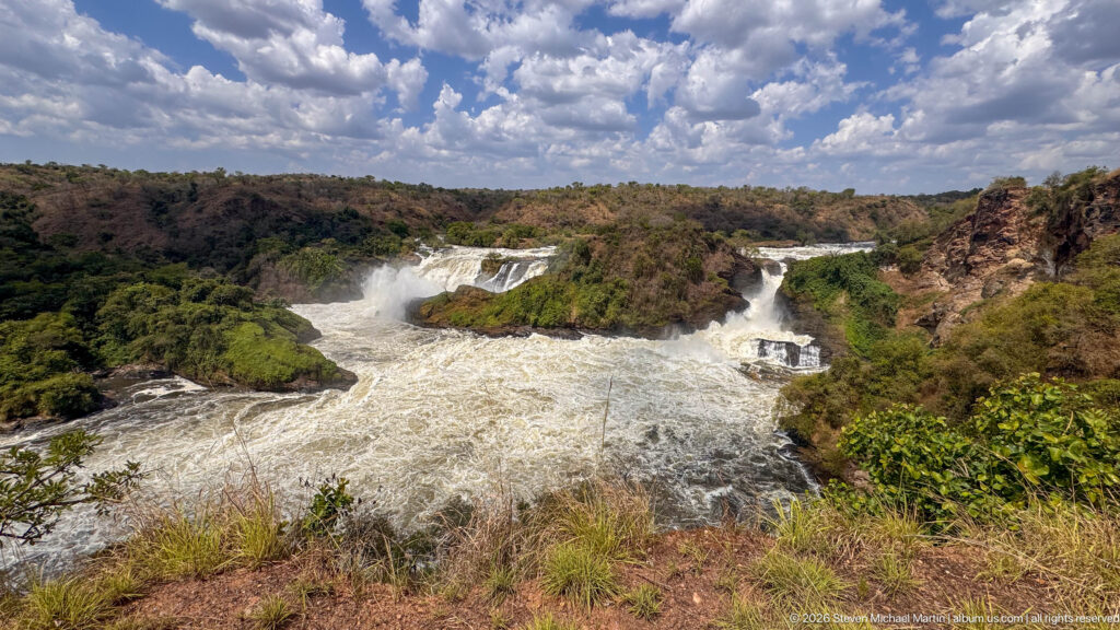 Wide elevated view of Murchison Falls, whitewater channels, green rock islands, dry hills, and cloudy blue sky.