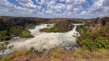 Wide elevated view of Murchison Falls, whitewater channels, green rock islands, dry hills, and cloudy blue sky.