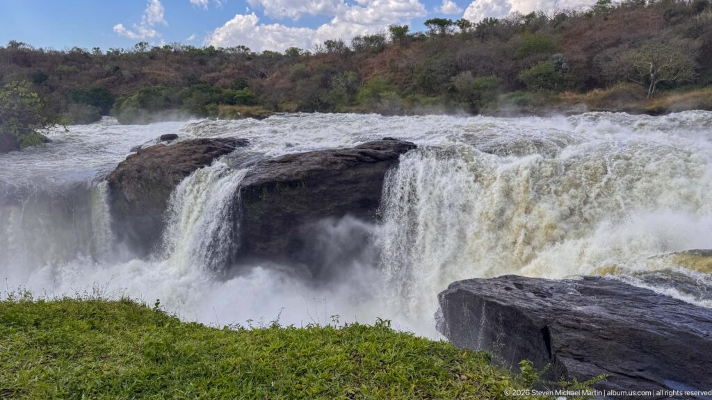 Heavy whitewater dropping in several curtains over dark bedrock at Murchison Falls, with mist rising below.