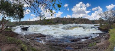 Broad whitewater rapids flowing over dark rock ledges above Murchison Falls beneath a blue, cloud-filled sky.