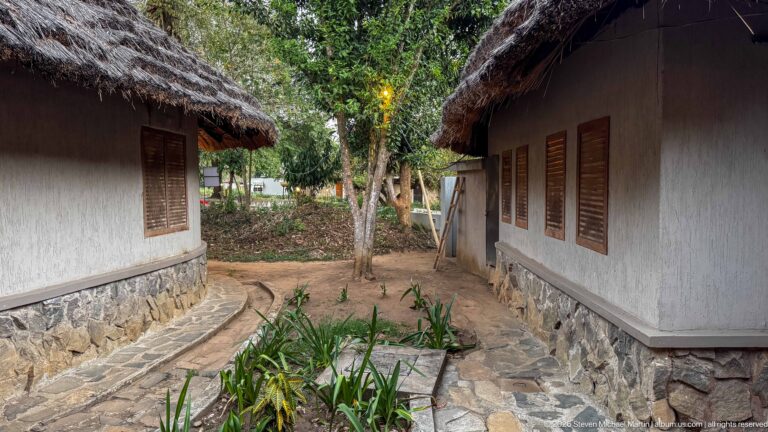 Narrow path between pale lodge buildings with shuttered windows, stone foundations, and deep thatched eaves.