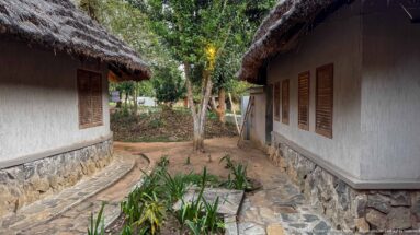 Narrow path between pale lodge buildings with shuttered windows, stone foundations, and deep thatched eaves.