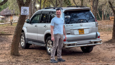 Man standing beside a silver Toyota Land Cruiser in a dry, wooded parking area.