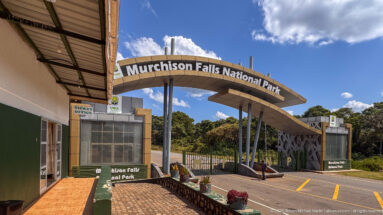 Oblique view of Murchison Falls National Park entrance with ticket office, curved gateway, road lanes, and security gate.