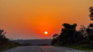 Orange sun setting above a paved road, with dark trees and distant low hills on the horizon.