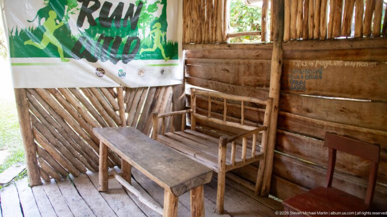 Rustic timber room with low wooden table, bench, chairs, slatted walls, open window, and a hanging banner.