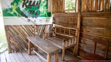Rustic timber room with low wooden table, bench, chairs, slatted walls, open window, and a hanging banner.