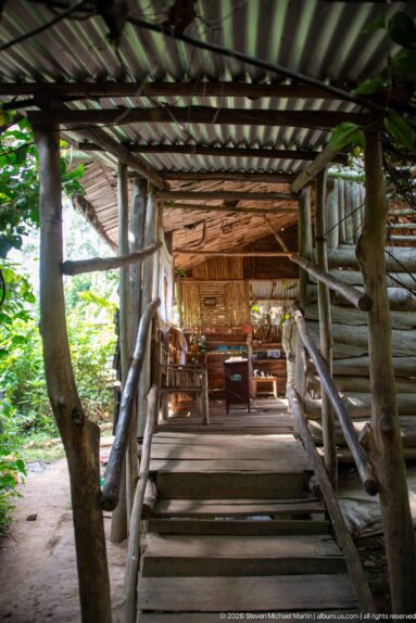 Steps and rustic branch rails leading into an open-air pavilion with wooden chairs, tables, and a corrugated roof.