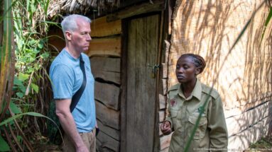 Uniformed staff member speaking with a visitor beside a rustic building of timber planks and woven wall panels.
