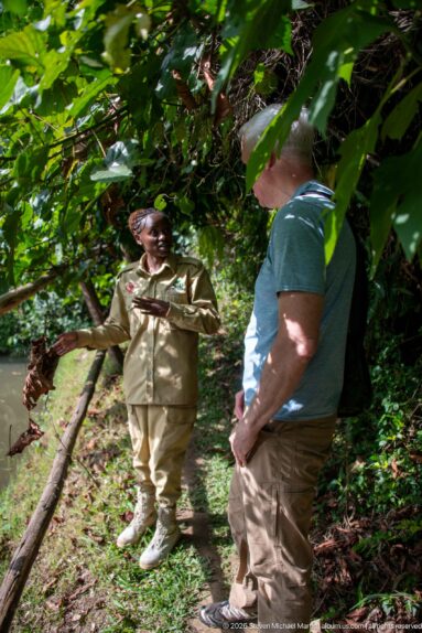 Uniformed staff member gesturing while speaking with two visitors beside a rustic rail on a shaded forest path.