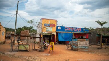 Blue-and-red Comfoam-branded roadside shop beside timber kiosks, a bicycle, brick walls, and scattered goods.