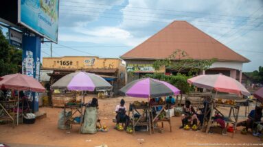 Several colorful umbrellas shading fruit stalls and seated vendors across a red-earth market area in front of roadside shops.