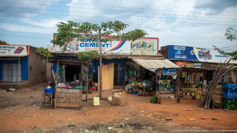 Open-front roadside shops beneath cement and motorcycle advertising with produce, containers, timber poles, and a bicycle outside.