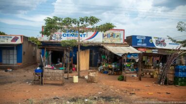 Open-front roadside shops beneath cement and motorcycle advertising with produce, containers, timber poles, and a bicycle outside.