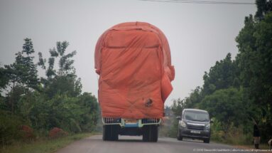 Freight truck carrying a very tall reddish tarpaulin-covered load on a two-lane road with an oncoming van.