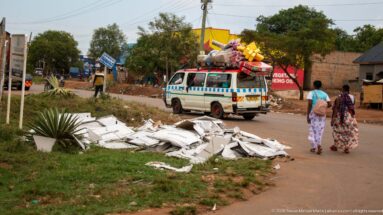 White-and-blue minibus carrying a tall roof load of household goods as two women walk along the town road.