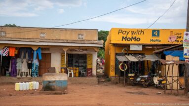 Yellow MoMo mobile-money shop beside a clothing storefront, parked motorcycles, mannequins, and jerrycans on a dirt frontage.