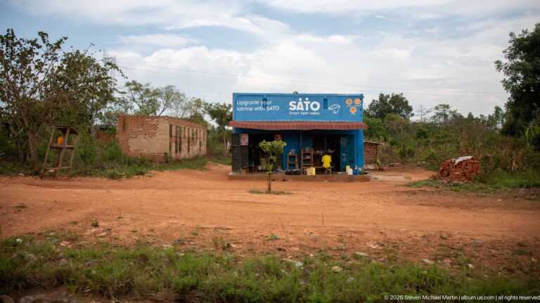 Small blue SATO-branded roadside kiosk with plastic chairs beside a red-earth road and sparse surrounding development.