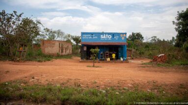 Small blue SATO-branded roadside kiosk with plastic chairs beside a red-earth road and sparse surrounding development.