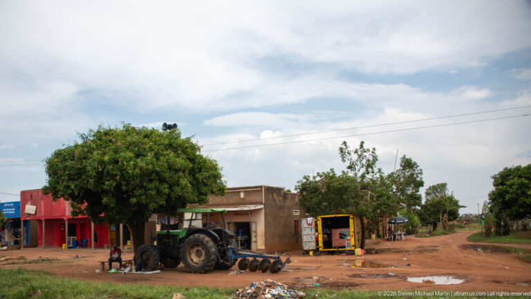 Large tractor parked on a red-earth roadside beside small shops, a kiosk, trees, and scattered machinery.