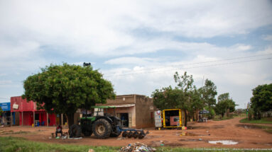 Large tractor parked on a red-earth roadside beside small shops, a kiosk, trees, and scattered machinery.