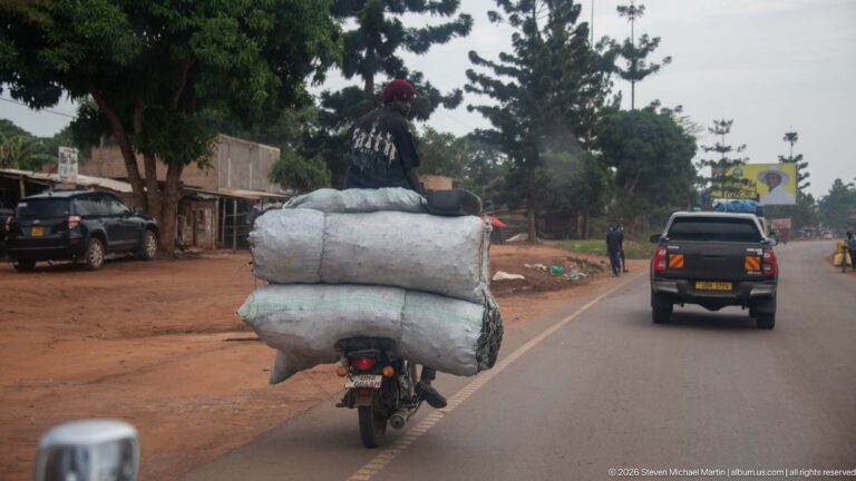Rear view of a motorcycle carrying huge white sacks while following vehicles along a paved town road.
