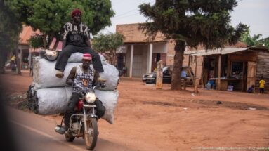 Motorcyclist approaching with enormous white sacks strapped around the bike and another person seated high on the load.