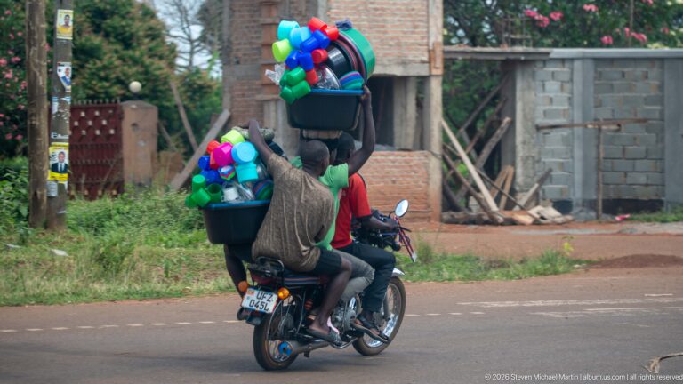 Motorcycle carrying brightly colored plastic basins, cups, buckets, and containers stacked high behind two riders.