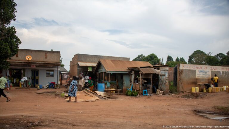 Row of simple timber, metal, and masonry shops along a dirt road with people walking between open-front businesses.